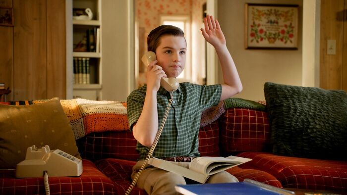 Young Sheldon child star sitting on a couch, holding a phone, and raising his hand in a cozy living room setting.