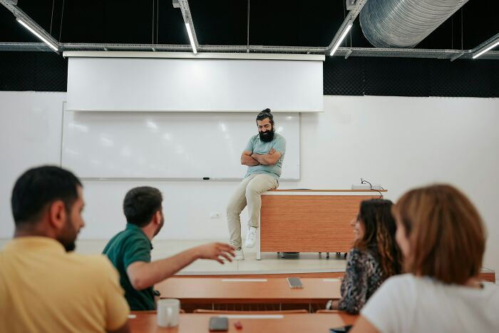 A man with a beard sitting on a desk in a classroom, appearing arrogant while others are talking in front of him.