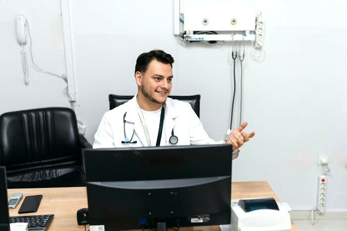 Male doctor with stethoscope smiling and gesturing while sitting at a desk in a medical office environment.