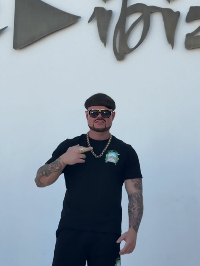 Man with distinctive haircut and beard wearing sunglasses and gold chain, posing in front of Ibiza sign, Ibiza Final Boss style. Man with distinctive haircut and beard wearing sunglasses and gold chain, posing in front of Ibiza sign, Ibiza Final Boss style.