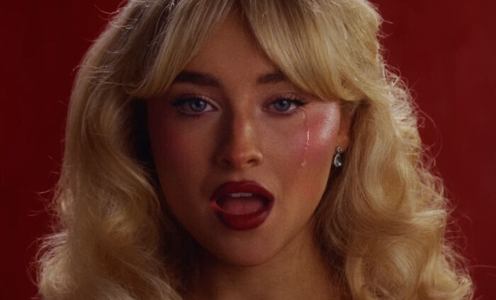 Sabrina Carpenter with blonde hair and red lipstick, posing against a red background in an expressive close-up shot.