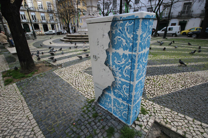 Street art on a public utility box designed to look like blue and white decorative tiles in an urban park setting.