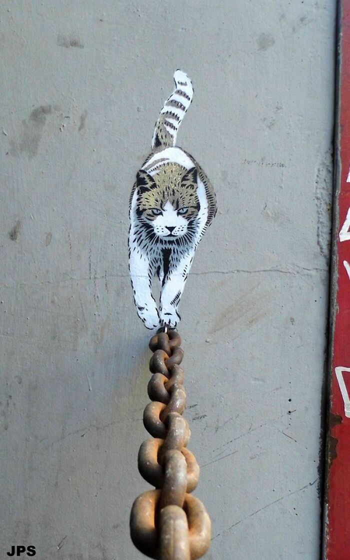 Street art of a cat walking on a rusty chain painted on a gray wall blending with the environment.