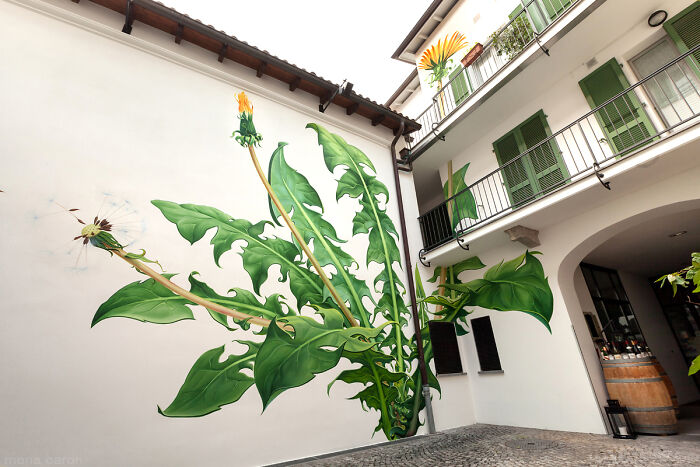 Large detailed street art mural of green leaves and a bird on a white building wall in an urban courtyard scene.
