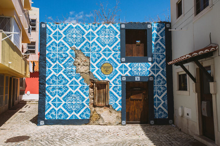 Blue and white patterned street art painted on an old building wall with wooden door and window in a sunny alley.