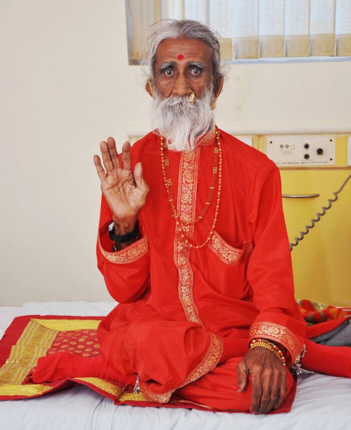 Elderly man in traditional red attire meditating and gesturing, showcasing traits of superhumans walking among us.