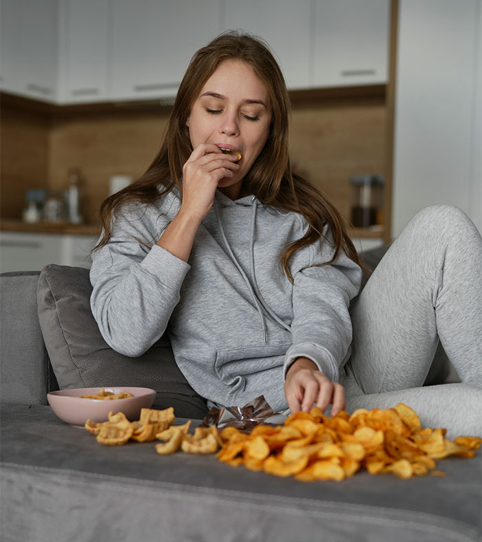 Young nanny from abroad eating snacks while sitting on a couch, reflecting American family houses food restrictions.