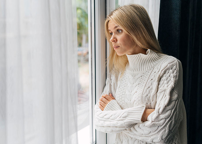Woman in a cozy white sweater looking thoughtfully out the window, reflecting on petty revenge against entitled dog owner.