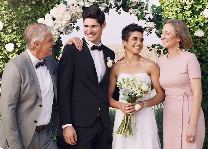 Bride in white dress holding bouquet standing with groom and parents under floral arch, illustrating woman refuses to hand over savings.