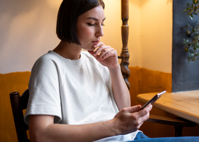 Woman in white shirt thoughtfully looking at phone, facing dilemma over savings after parents claim financial rights.
