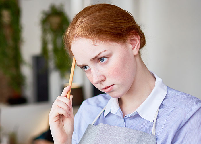 Young woman in apron holding pencil to head, looking thoughtfully concerned in an indoor pet store setting.