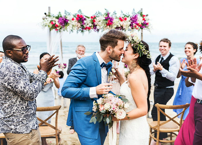 Couple kissing at a beach wedding ceremony while guests applaud, illustrating social dynamics and coworker relationships.