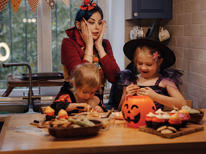 Mom refusing to leave kids at home for Halloween party, stressed woman watching children in festive costumes at kitchen table. - 1