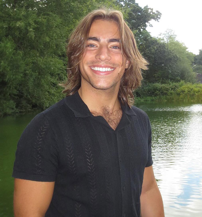 Chase Filandro smiling outdoors near a lake, wearing a black shirt with long hair and greenery in the background.
