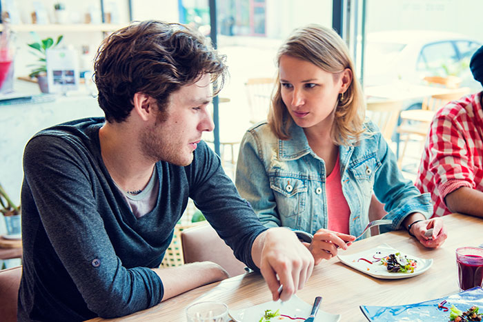 Man upset as girlfriend eats peanut butter cake at work, knowing he cannot eat it, showing frustration during lunch.