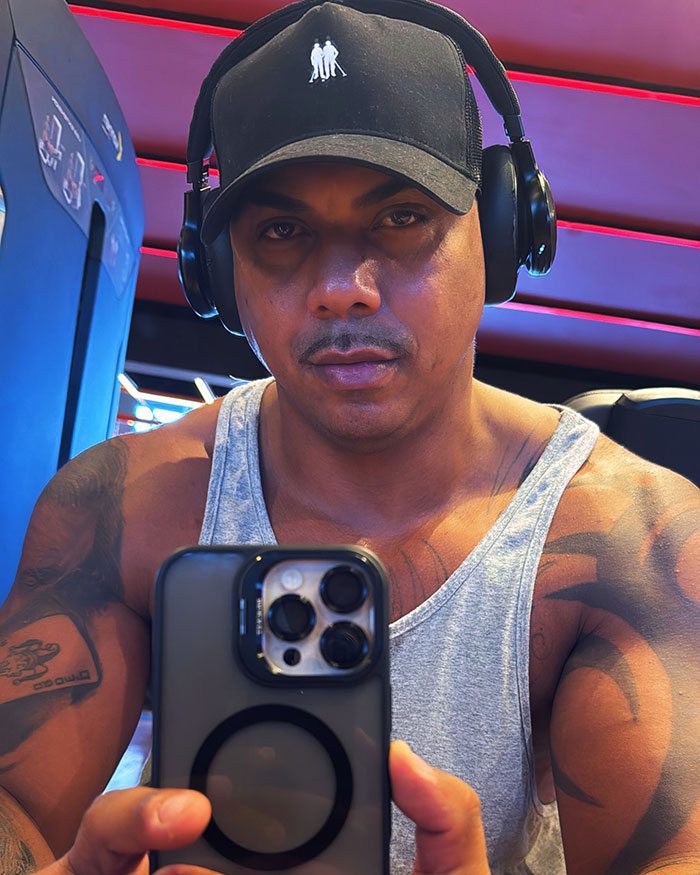Man with headphones and black cap taking a selfie in gym, showcasing muscular arms and tattoo in a sleeveless gray shirt.