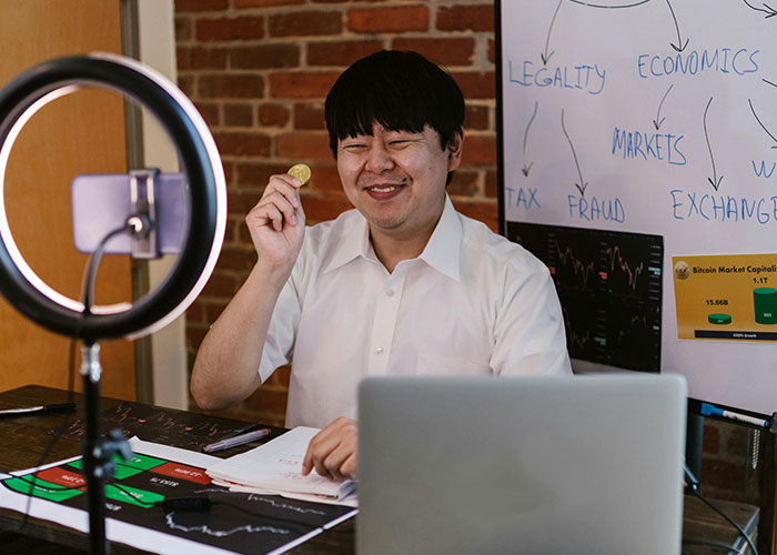 Man holding a coin in front of laptop with Bitcoin charts, illustrating jobs related to economics and markets without a soul.