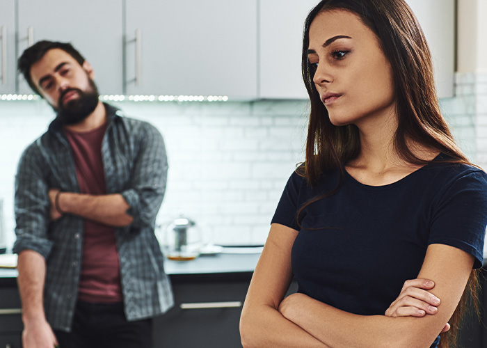 A young woman ignoring her gut feeling stands upset while a man watches her in a kitchen, highlighting regret and luck to be alive.