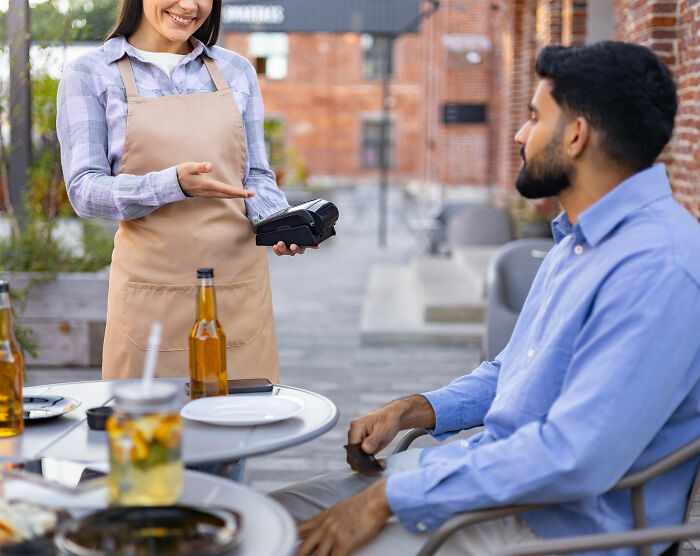 Waitress smiling and holding a card reader while a man in a blue shirt sits at an outdoor table with drinks.