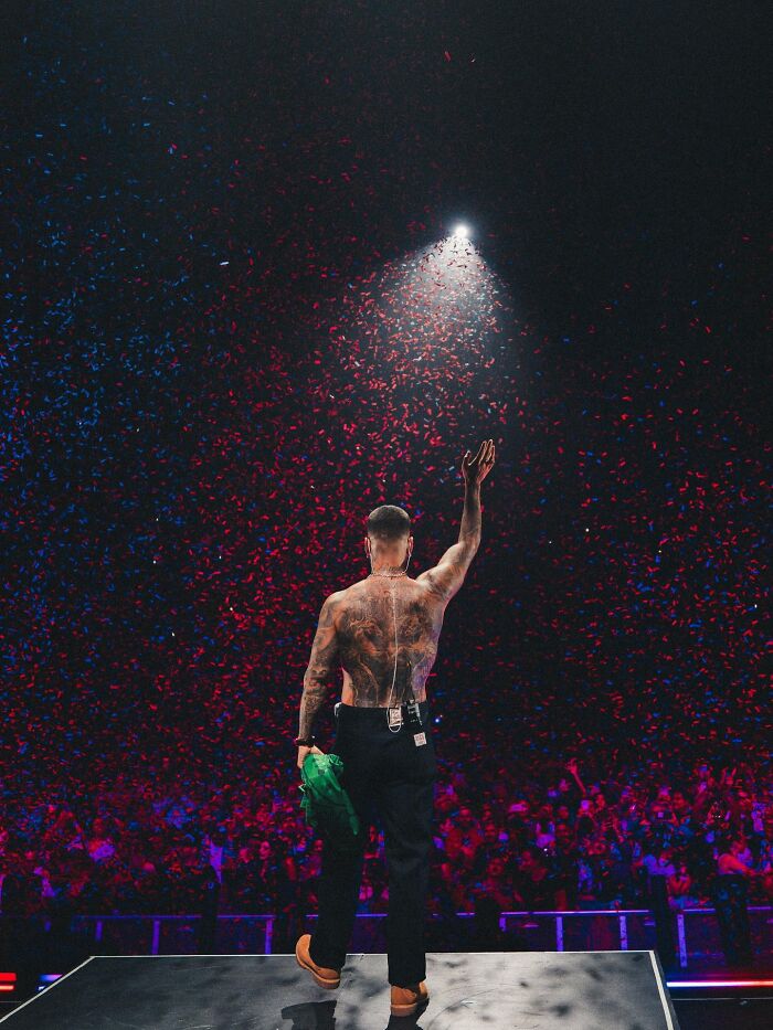 Singer on stage with tattoos, raising hand amid red and blue confetti during concert abruptly stopped for irresponsible mom. Singer on stage with tattoos, raising hand amid red and blue confetti during concert abruptly stopped for irresponsible mom.