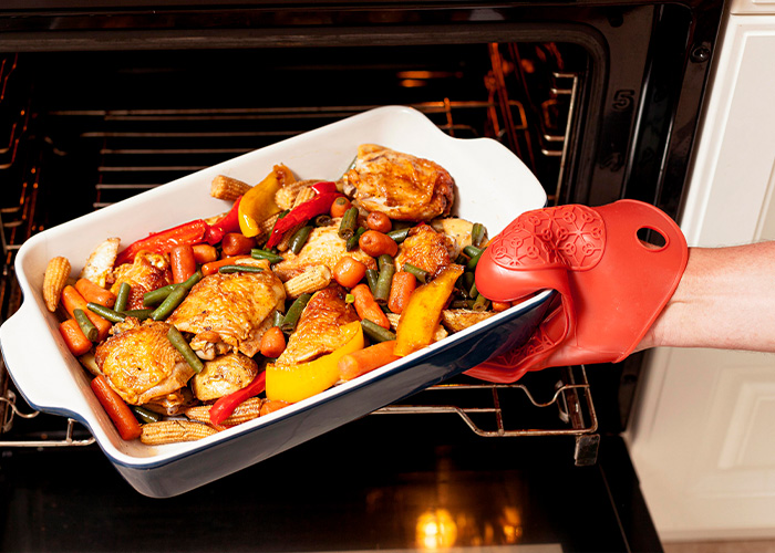 Person wearing red oven mitt removing a tray of roasted chicken and vegetables from an oven in a kitchen setting.