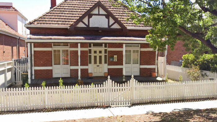Historic abandoned million-dollar house with white picket fence, surrounded by trees and neighboring homes.