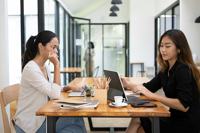 Two women working on laptops at a wooden table in a modern office, one looks thoughtful while the other types.