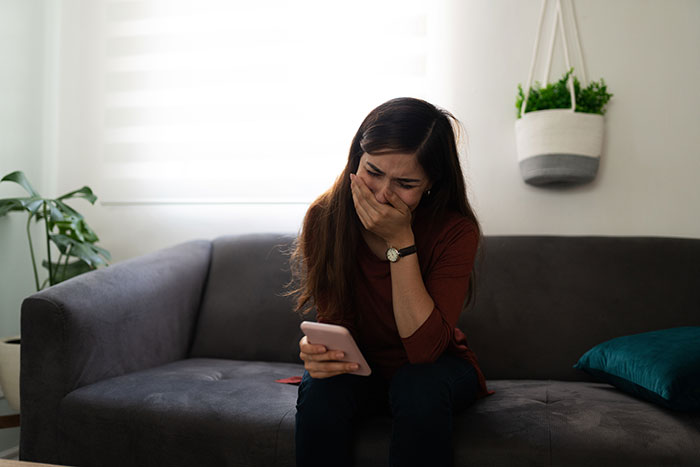 Sad woman sitting on couch, covering mouth while reading distressing message on phone about euthanizing dog online controversy