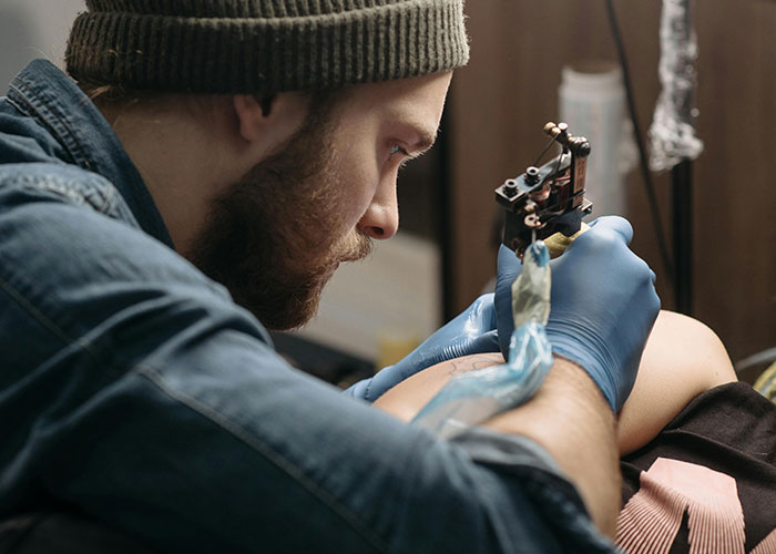 Tattoo artist wearing a beanie and blue gloves carefully working on a client's arm in a focused indoor setting.