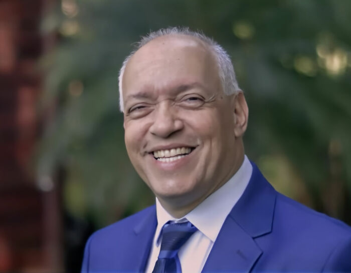 Smiling pastor in a blue suit and tie, pictured outdoors with blurred greenery in the background. Smiling pastor in a blue suit and tie, pictured outdoors with blurred greenery in the background.