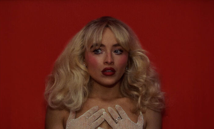 Blonde woman with dramatic makeup and fishnet gloves poses against a red background, evoking a sensual pole dance vibe.