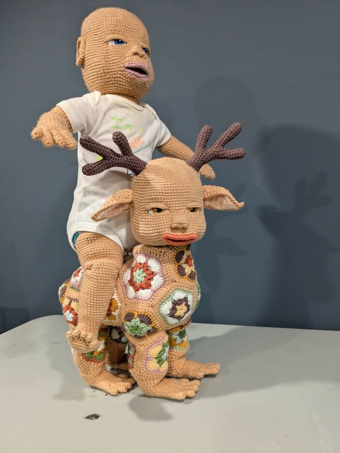 Crocheted bizarre humanoid doll riding a multicolored crochet creature with antlers in a craft shaming DIY project.