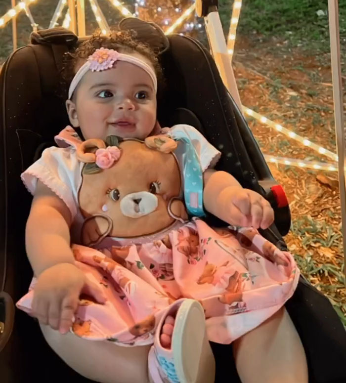 Toddler sitting in car seat wearing pink dress with teddy bear design, surrounded by festive outdoor lights and greenery.