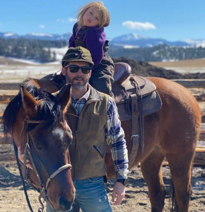 Man standing beside horse with child on saddle in a rural landscape, related to Brandon Blackstock's obituary mention.