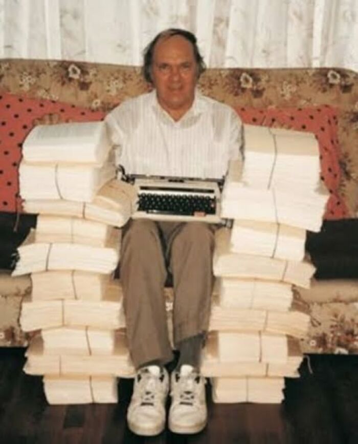 Man sitting among large stacks of paper with a typewriter, illustrating unsettling images from morbid knowledge.