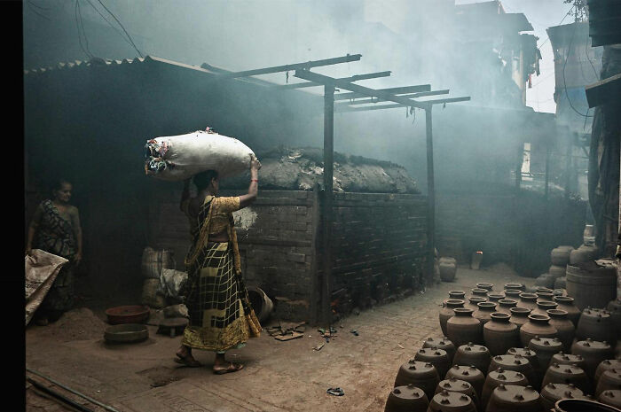Woman carrying a large sack on her head in a smoky pottery workshop, showcasing candid everyday moments from Asia.