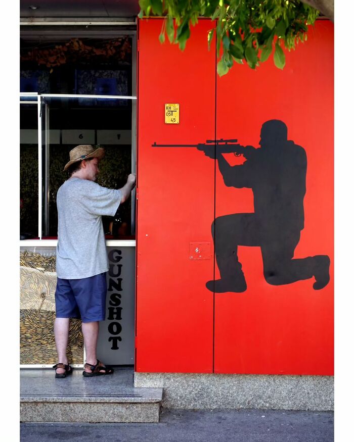 Man in hat standing next to red wall with black silhouette of kneeling figure aiming a rifle in unseen street moment.