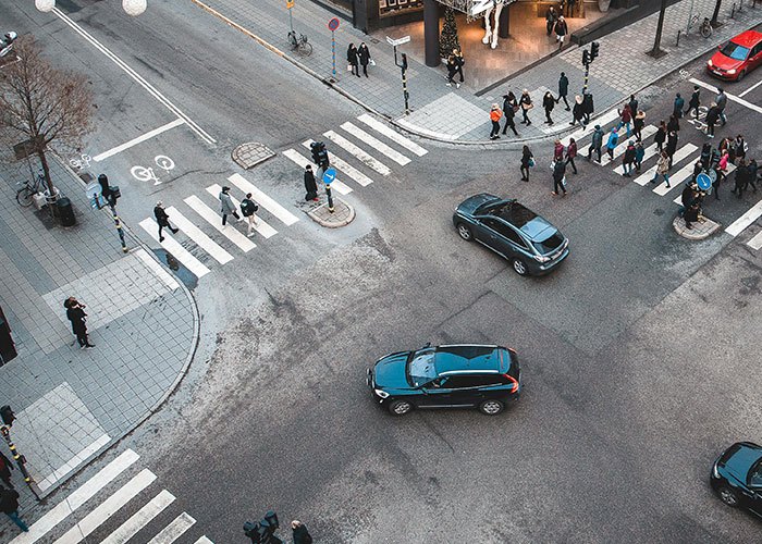 Cars driving over the speed limit at a busy city intersection with pedestrians crossing and waiting on sidewalks.