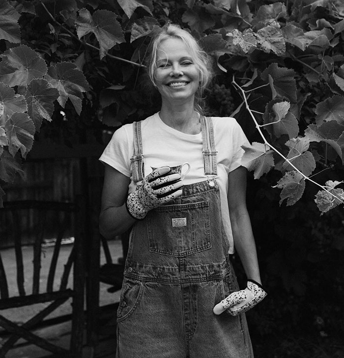 Pamela Anderson smiling outdoors in denim overalls and gardening gloves amid grapevine leaves, addressing cooking show controversy. - 14