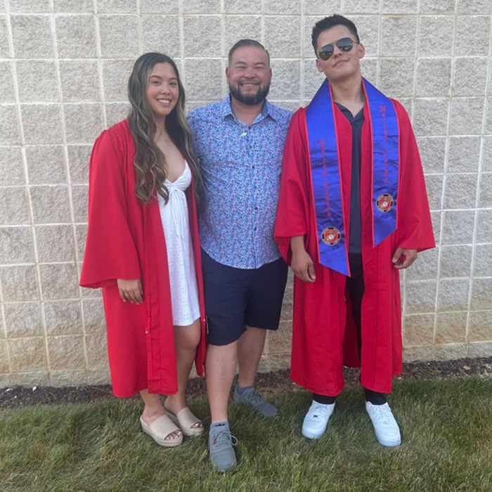 Kate and Jon Gosselin&rsquo;s son Collin standing with family, wearing red graduation gown and blue sash outdoors near a brick wall.