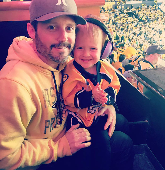 Man in a cap and hoodie holding a smiling child wearing earmuffs and a hockey jersey at a crowded sports event.