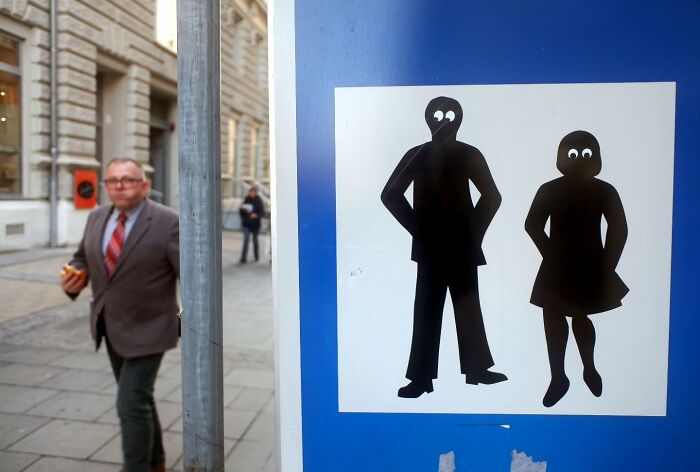 Man in a suit holding a sandwich walking on the street near a blue sign with cartoon figures with googly eyes.