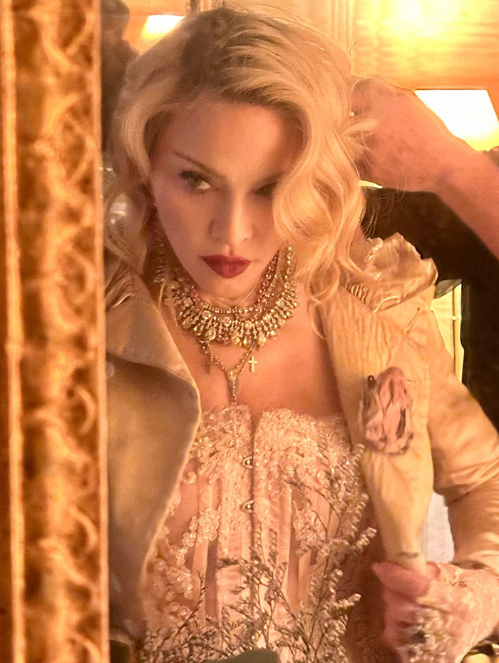 Blonde woman wearing elaborate jewelry and a lace outfit, reflecting in a mirror at an elegant indoor event.