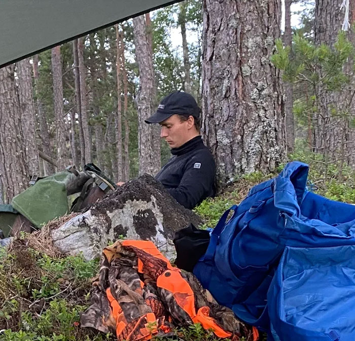 Young man resting against tree during hiking trip, surrounded by backpack and outdoor gear in forest setting.