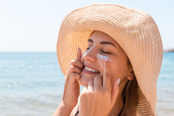Woman in a sun hat applying sunscreen on her face at the beach representing low effort high reward skin care tip.