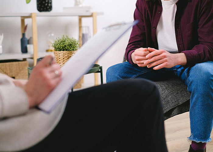 Person in casual clothes sitting on a couch, hands clasped, during a conversation with another holding a clipboard.