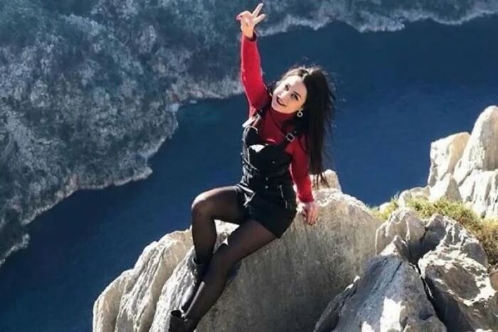 Young woman in a black dress and red top posing on rocky cliff edge with lake below, unsettling image from Morbid Knowledge group