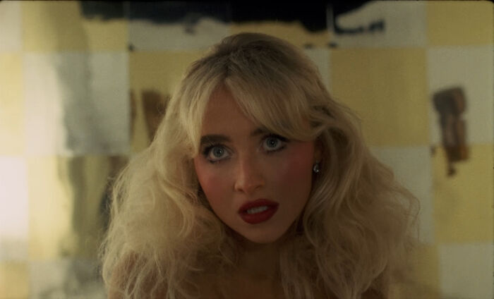 Blonde woman with voluminous hair and red lipstick looks intently in a dimly lit room, related to pole dance video content.