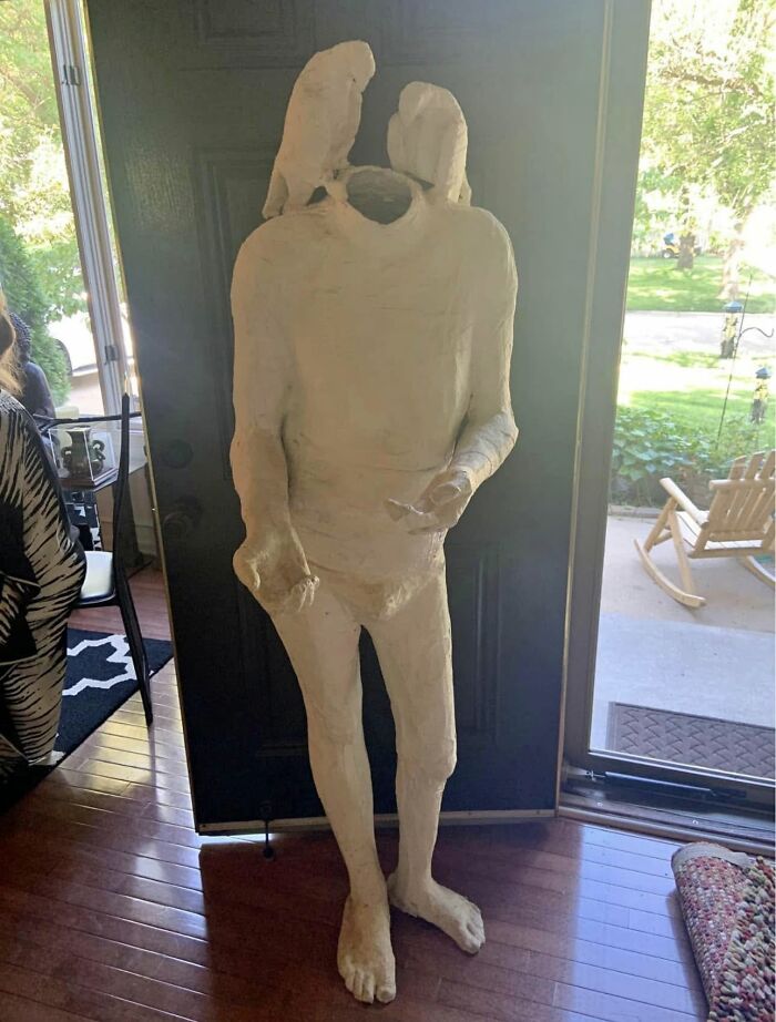 Awkward DIY craft sculpture of a headless human figure with rough texture standing indoors by a door.