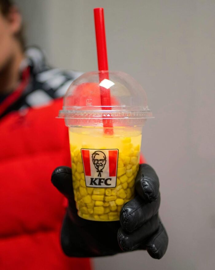 KFC plastic cup filled with corn kernels and a red straw held by a gloved hand, highlighting unhealthy food choices.